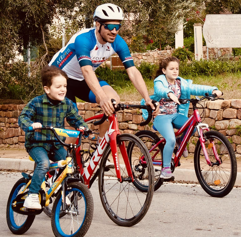 Photo of Greek road and track cyclist Polychronis_Tzortzakis riding with 2 children and wearing the Acropolis Organics logo on his shirt, showing the benefits of healthy foods for healthy living.
